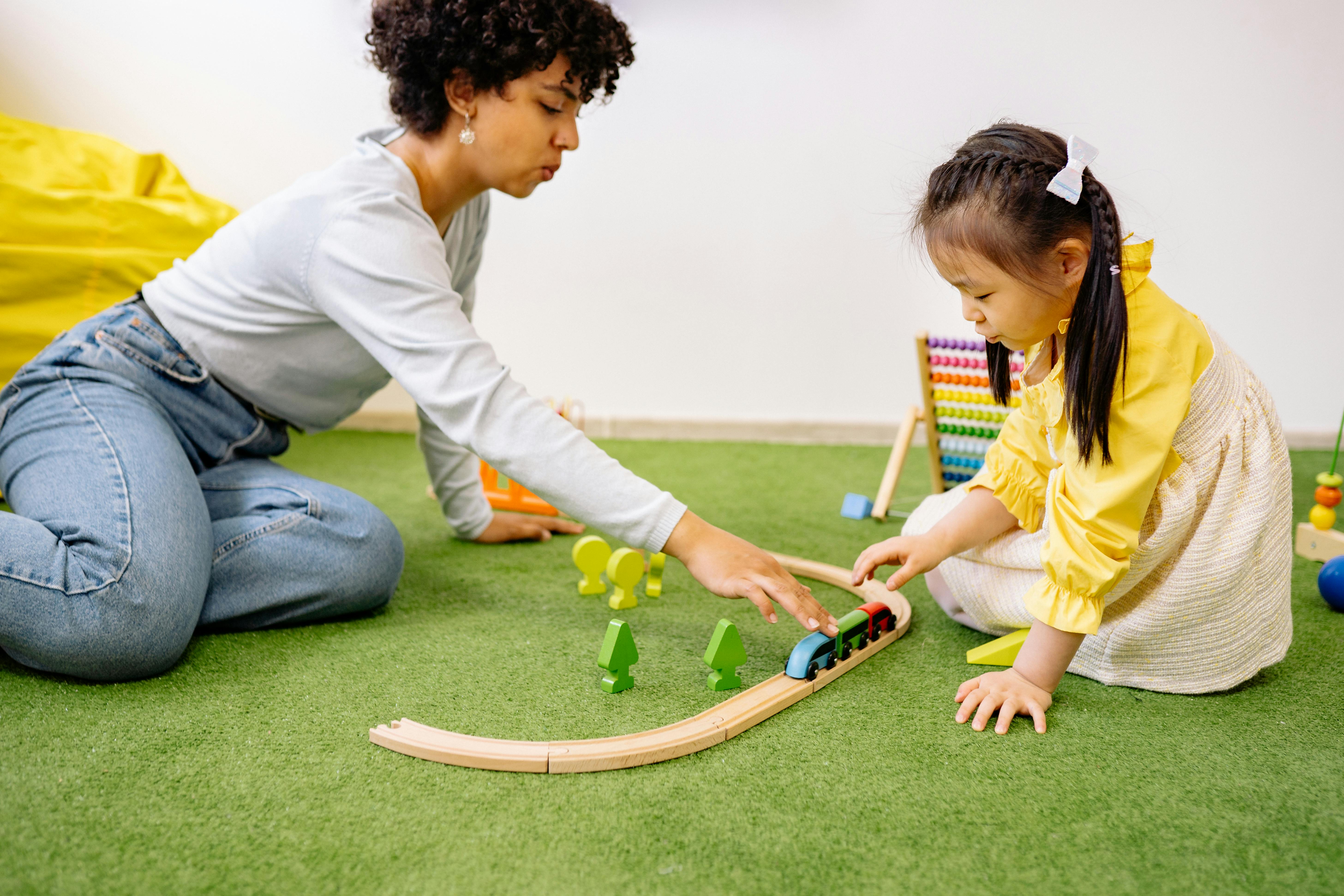 An instructor and a child are seated on the floor playing with a wooden train.