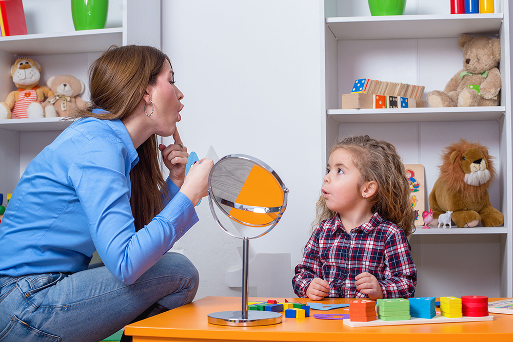 A woman wearing a blue shirt and who has long hair is kneeling and gesturing to a young child looking at up at her from a table with colored blocks. 