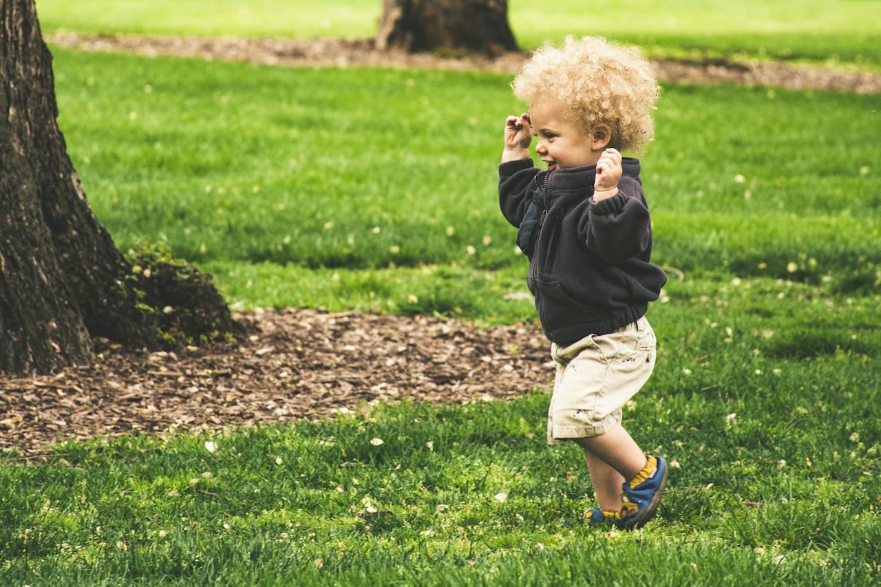 "A child with curly blond hair smiles while running outside in a park setting"
